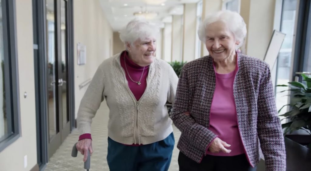 Two older adults walk through the halls of a retirement home, smiling and enjoying each other's company.
