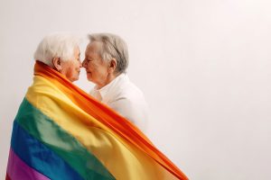Two older women close to each other, nose touching. A rainbow flag is draped around both of them.