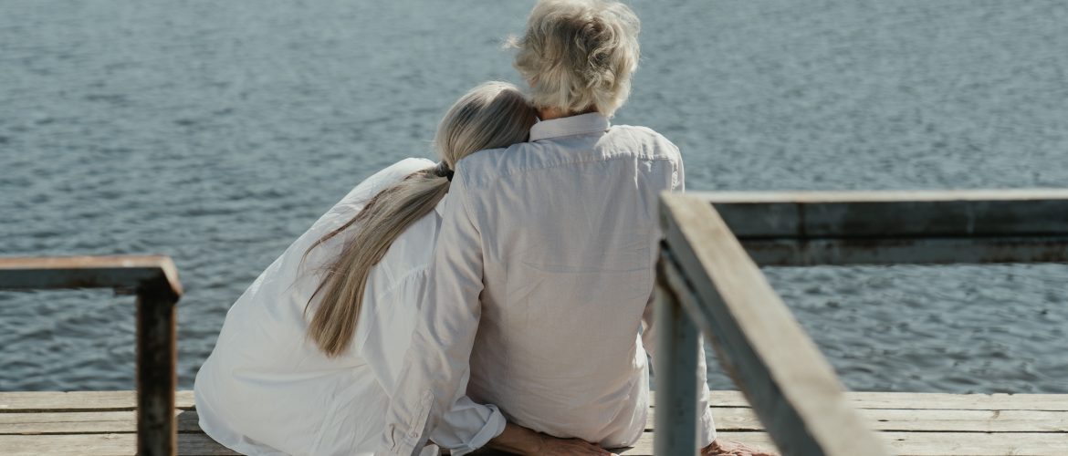Older couple snuggling on a dock in front of water, their backs to the camera.