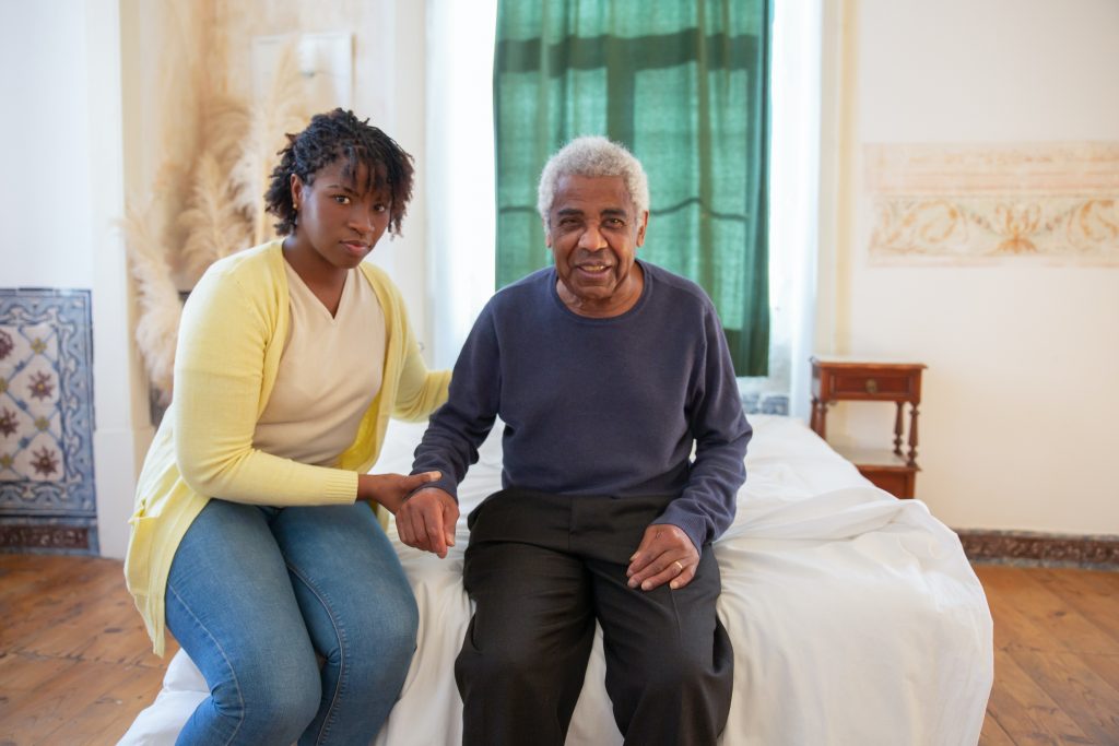 White-haired man and younger female caregiver sit on bed, caregiver steadying man's wrist