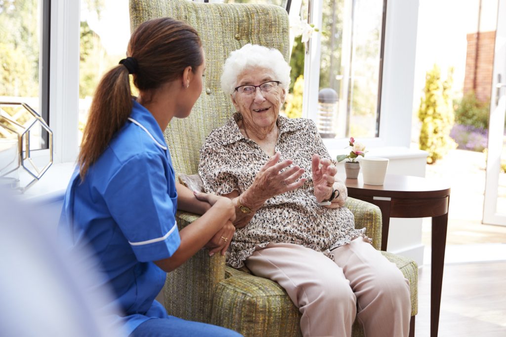 Senior Woman Sitting In Chair And Talking With Nurse In Retirement Home