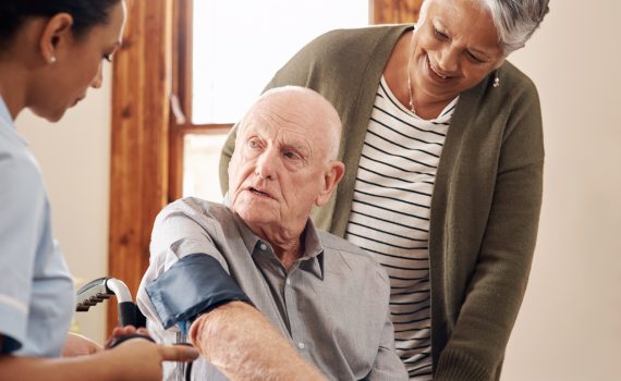 Senior man uses blood pressure cuff while point-of-care worker and senior woman look on